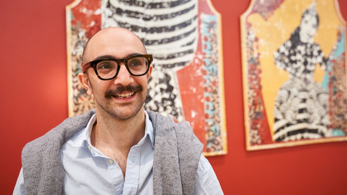 A man with a moustache and glasses smiles in front of a deep red wall with two large prints hanging on it