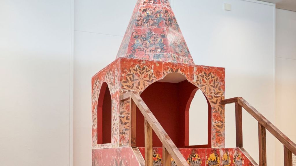 the top of a square tower structure with a pointed cap on top, with a balustrade and stairs leading up to it, all covered in intricate prints in red, ochre and black & white