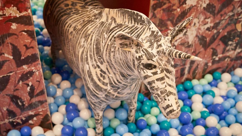 A sculpture of a zebra with a unicorn's horn on its head stands in a ball pit among balls of varied shades of blue